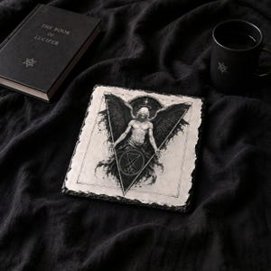 Puede incluir: Ilustraci&oacute;n en blanco y negro de un &aacute;ngel con grandes alas y un halo, sobre un posavasos cuadrado. Una taza negra y un libro titulado "THE BOOK OF LUCIFER" tambi&eacute;n est&aacute;n presentes en la imagen.