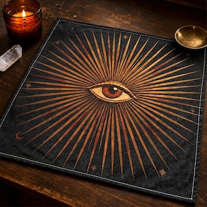 May include: A square black fabric cloth featuring a central graphic of a human eye surrounded by radiating golden-brown sunburst rays. The design includes small star icons and a crescent moon symbol, with white stitched edges on all four sides.