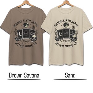 K&ouml;nnte beinhalten: Zwei kurz&auml;rmelige T-Shirts, eines braun und eines sandfarben, werden gezeigt. Jedes Shirt hat eine kreisf&ouml;rmige Grafik mit dem Text "MAMA'S SIXTH SENSE" und "WITCH MODE ON". Die Grafik enth&auml;lt Tarotkarten, einen Totenkopf und einen Mond.