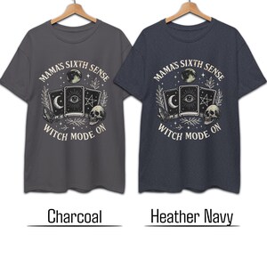 K&ouml;nnte beinhalten: Zwei T-Shirts, Holzkohle und Heather Navy, mit einem grafischen Design. Das Design enth&auml;lt den Text "MAMA'S SIXTH SENSE WITCH MODE ON" um einen Mond, Tarotkarten, einen Sch&auml;del und andere mystische Elemente. Die Hemden h&auml;ngen an einem Holzkleiderb&uuml;gel.
