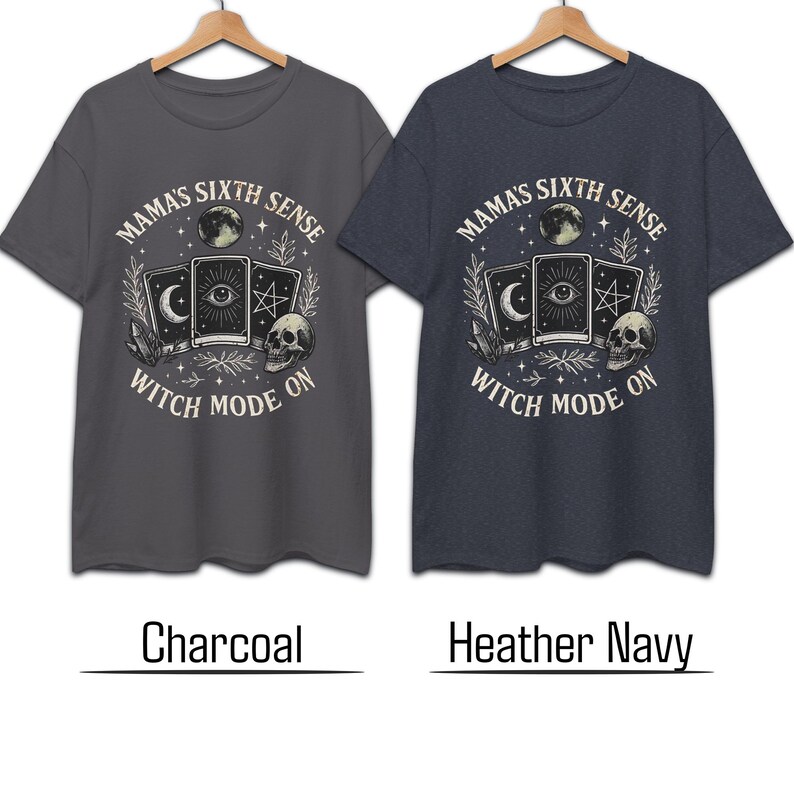 K&ouml;nnte beinhalten: Zwei T-Shirts, Holzkohle und Heather Navy, mit einem grafischen Design. Das Design enth&auml;lt den Text "MAMA'S SIXTH SENSE WITCH MODE ON" um einen Mond, Tarotkarten, einen Sch&auml;del und andere mystische Elemente. Die Hemden h&auml;ngen an einem Holzkleiderb&uuml;gel.