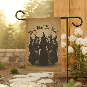 May include: A garden flag with the text "Girls Will Be Girls" above an illustration of five witches in black dresses and pointed hats, holding hands in a circle. The flag is displayed on a black metal stand.