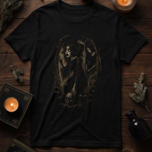May include: Black t-shirt with a sepia-toned illustration of two figures in a forest. One figure reaches towards a skull, the other stands behind. The artwork has a gothic, horror theme.