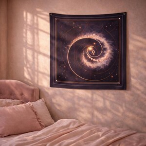 May include: A dark blue tapestry with a golden spiral galaxy design, featuring a central symbol and numerous stars. The tapestry is square with a stitched border and grommets in each corner. The tapestry is hanging on a wall above a bed with pink bedding.