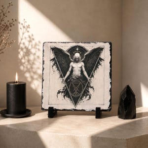 Puede incluir: Impresi&oacute;n en blanco y negro de un &aacute;ngel con grandes alas, sobre una piedra cuadrada. La obra de arte se exhibe en un soporte negro. Una vela negra y un cristal negro est&aacute;n cerca.