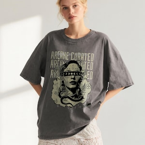 May include: Gray oversized t-shirt with the text "ARCANE CURATED" stacked vertically. The graphic features a woman's face with a snake and moon phases. The shirt has short sleeves and a crew neck.