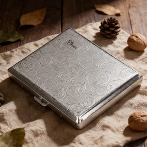 Könnte beinhalten: Eine silberne Zigarettenetui mit einem eingravierten floralen Muster. Der Name "Ethan" ist oben eingraviert. Die rechteckige Hülle hat einen Verschluss. Der Hintergrund umfasst Walnüsse, einen Tannenzapfen und Blätter.
