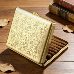 Peut inclure: Étui à cigarettes en métal doré, ouvert pour révéler plusieurs cigares bruns. L'étui présente un motif floral en relief orné. L'étui rectangulaire est doté d'un fermoir doré. L'arrière-plan comprend des livres et une surface en bois.