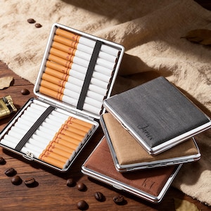 Könnte beinhalten: Eine Auswahl an Zigarettenetuis in verschiedenen Farben, darunter Grau, Beige und Braun, mit silbernen Metallkanten. Geöffnete Etuis zeigen Zigaretten, die von schwarzen Gummibändern gehalten werden. Ein Etui ist mit dem Namen "James" graviert.