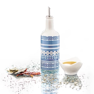 Può includere: Una bottiglia di olio d'oliva in ceramica bianca con un motivo geometrico blu e beige. La bottiglia ha un beccuccio di versamento argentato ed è appoggiata su una superficie bianca con una piccola ciotola bianca piena di olio e spezie sparse.