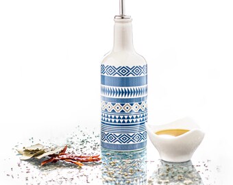 Puede incluir: Una botella de aceite de oliva de cerámica blanca con un patrón geométrico azul y beige. La botella tiene un pico de vertido plateado y está sobre una superficie blanca con un pequeño cuenco blanco lleno de aceite y especias esparcidas.
