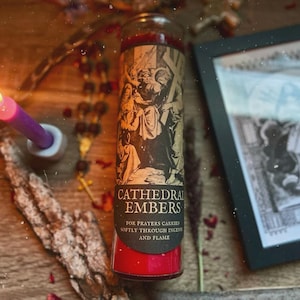 Puede incluir: Una vela roja en un frasco de vidrio con una etiqueta que presenta una ilustración religiosa y las palabras "Cathedral Embers". La vela está rodeada de otros elementos espirituales, como un rosario y una imagen enmarcada.
