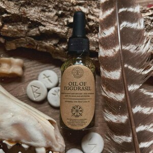 May include: A small glass bottle of "Oil of Yggdrasil" with a black dropper, set against a backdrop of wood, runes, and a feather. The label is brown with white text. The bottle contains a golden liquid. The text on the label reads "External Use Only 1 fl oz."