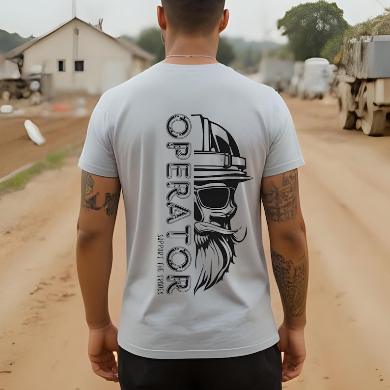 Man wearing a light gray t-shirt with a bold vertical graphic on the back that says "OPERATOR" with a bearded skull wearing a hard hat and sunglasses; below it reads "Support the Trades." The shirt is designed for heavy equipment operators and construction workers, photographed on a dirt road jobsite for a rugged, blue-collar vibe.
