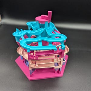 May include: A pink and blue 3D printed marble run toy with a hexagonal base. The toy has multiple levels and a winding track for marbles to travel through.