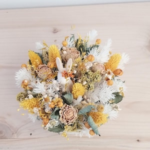 Peut inclure: Une composition florale circulaire avec une figurine de lapin blanc parmi des fleurs séchées. L'arrangement comprend des fleurs jaunes, blanches et beiges, avec des accents de feuillage vert. Le lapin a des accents orange. L'esthétique générale est rustique et naturelle.