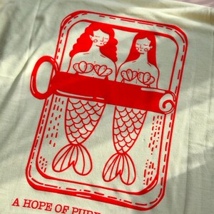 Op de afbeelding: Crèmekleurig T-shirt met een rode afbeelding van twee zeemeerminnen in een sardineblik. Het ontwerp bevat de tekst "A HOPE OF PURE FREEDOME". De zeemeerminnen hebben lang rood haar en schelpen tops.