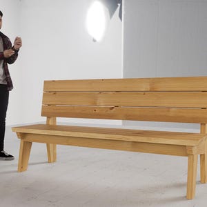 May include: A wooden bench with a backrest made of horizontal planks. The bench is constructed from light-colored wood and has a simple, modern design. The bench is approximately 60 inches long.