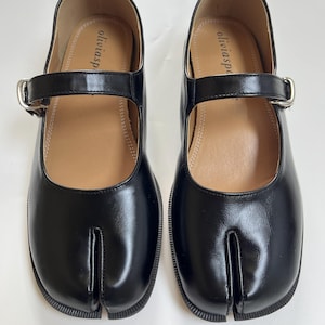 May include: A pair of black leather Tabi shoes with a split toe design. The shoes feature a Mary Jane style strap with a silver buckle. The soles are black, and the insoles are tan. The brand name "oliviasp" is printed on the insole.