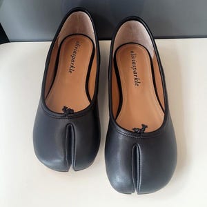 Può includere: Un paio di scarpe basse nere con punta divisa, finitura liscia e un piccolo fiocco nero sul davanti. Le solette interne color cuoio riportano il nome del marchio oliviasparkle stampato in nero. Le scarpe presentano un caratteristico design a fessura.