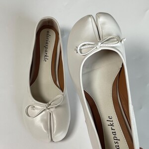 May include: A pair of white split-toe ballet flats featuring a distinctive cleft design at the front, a small decorative bow on the vamp, and a tan interior insole printed with the brand name oliviasparkle in black lowercase lettering.