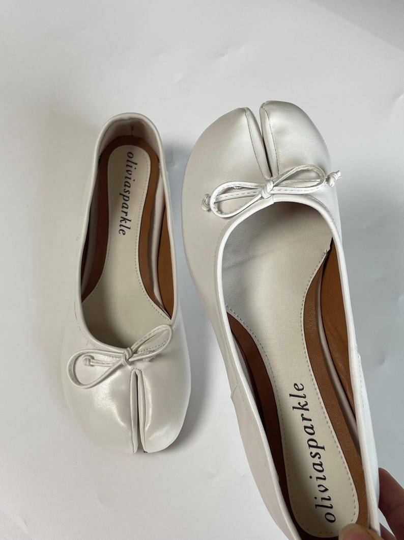May include: A pair of white split-toe ballet flats featuring a distinctive cleft design at the front, a small decorative bow on the vamp, and a tan interior insole printed with the brand name oliviasparkle in black lowercase lettering.
