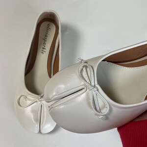 May include: A pair of white split-toe ballet flats featuring a distinctive cleft design at the front, a small decorative bow on the vamp, and a tan insole with the brand name oliviasparkle printed in black text on the heel area of the shoe.