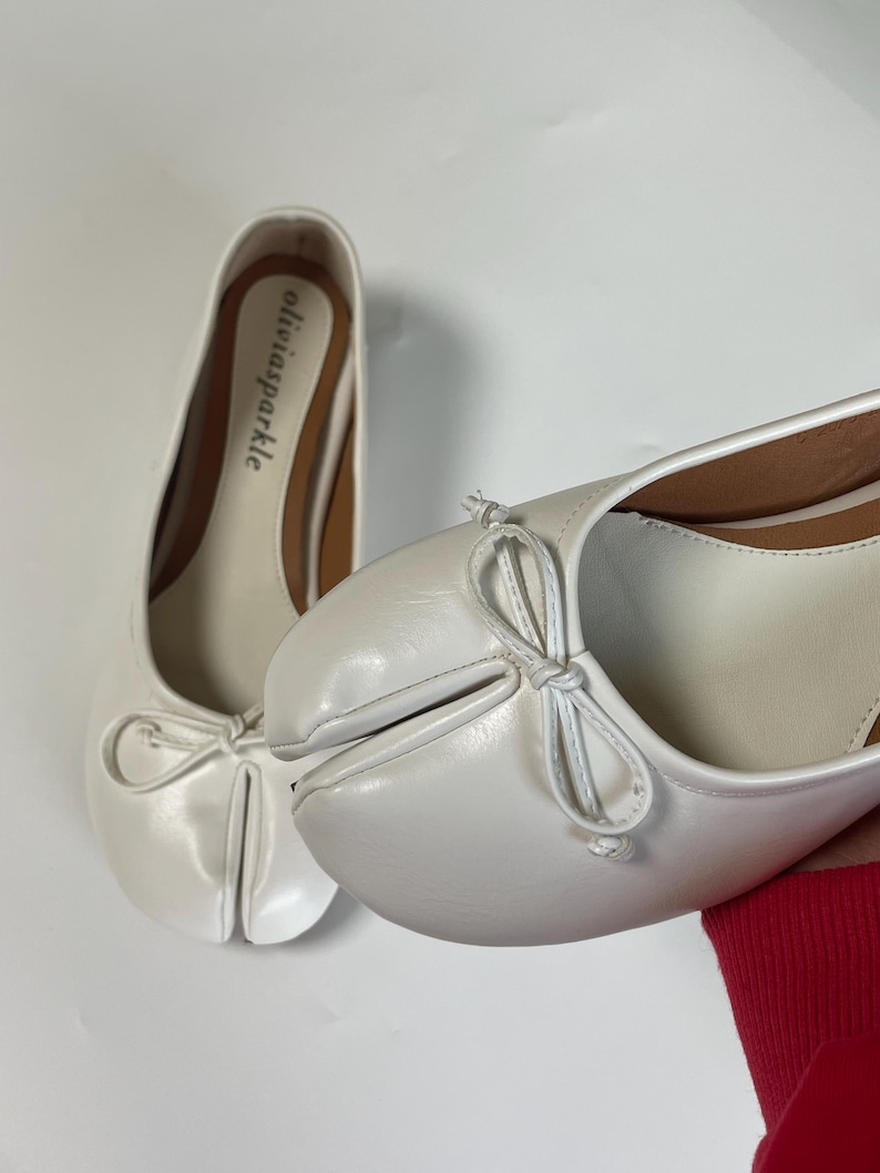 May include: A pair of white split-toe ballet flats featuring a distinctive cleft design at the front, a small decorative bow on the vamp, and a tan insole with the brand name oliviasparkle printed in black text on the heel area of the shoe.