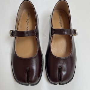 May include: A pair of dark brown split-toe Mary Jane shoes with a smooth finish, featuring a single strap across the instep with a silver-toned metal buckle. The interior insole is tan and displays the brand name oliviasparkle in black text.
