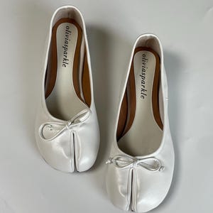 May include: A pair of white split-toe ballet flats featuring a small bow on the front and the brand name oliviasparkle printed in black on the inner sole. The shoes have a smooth finish and a distinct separation between the big toe and other toes.