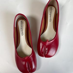 Può includere: Un paio di ballerine rosse con punta divisa e finitura liscia. Ogni scarpa presenta un piccolo fiocco rosso sulla tomaia e una soletta beige con il nome del marchio oliviasparkle stampato in nero. Il design include una punta separata.