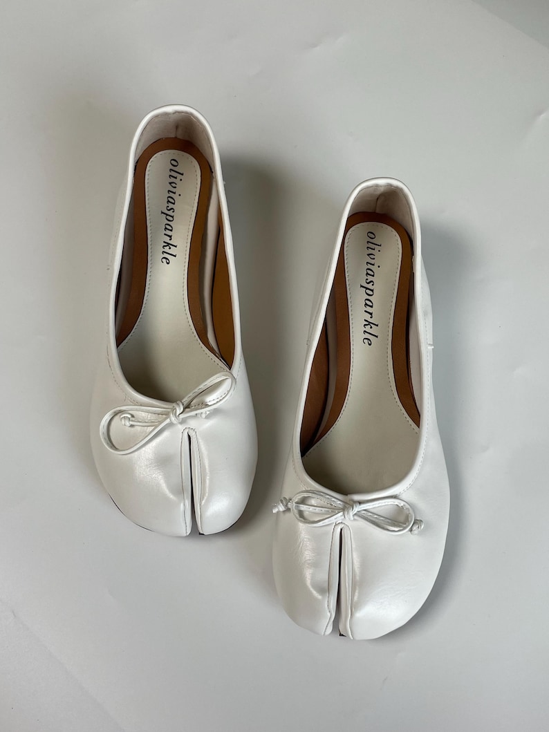 May include: A pair of white split-toe ballet flats featuring a small bow on the front and the brand name oliviasparkle printed in black on the inner sole. The shoes have a smooth finish and a distinct separation between the big toe and other toes.