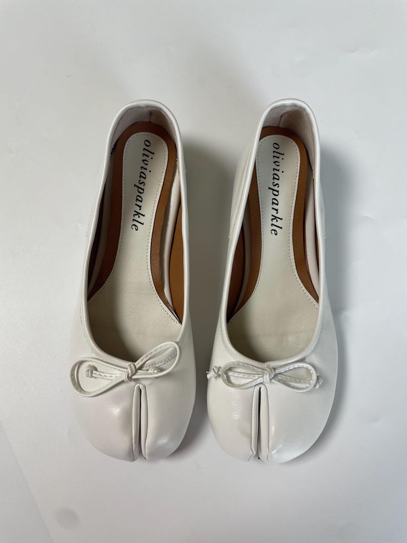 May include: A pair of white split-toe ballet flats featuring a small decorative bow on the front of each shoe. The interior footbed is tan with the brand name oliviasparkle printed in black text on the heel area of each shoe.