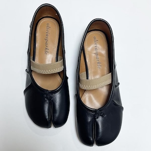 May include: A pair of black split-toe ballet flats featuring a beige elastic strap across the instep. The interior insole is tan and displays the brand name oliviasparkle in black text. The shoes have a rounded silhouette with a distinct toe split.