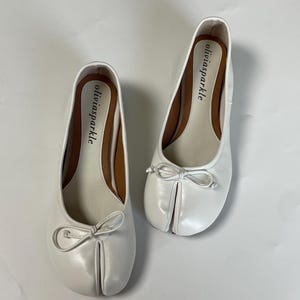 May include: A pair of white split-toe ballet flats featuring a small decorative bow on the front. The interior footbed is light beige with a brown trim and the brand name oliviasparkle printed in black text on the heel area of each shoe.
