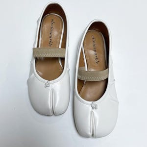 May include: A pair of white split-toe ballet flats featuring a beige elastic strap across the instep and a small white knot detail at the center front. The interior footbed is brown and displays the brand name oliviasparkle in black text.
