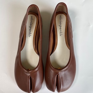 May include: A pair of brown split-toe flats featuring a distinct cleft design at the front. The interior insoles are cream-colored and feature the brand name oliviasparkle printed in black text. The shoes have a smooth finish and low profile.