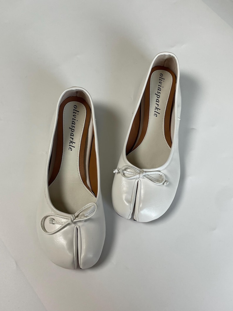 May include: A pair of white split-toe ballet flats featuring a small decorative bow on the front. The interior footbed is light beige with a brown trim and the brand name oliviasparkle printed in black text on the heel area of each shoe.