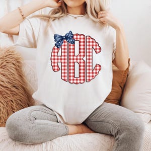 Op de afbeelding: Wit T-shirt met een rood-wit geruit monogram met de letters "ABC". Een blauwe strik met witte sterren siert de bovenkant van het monogram. Het T-shirt is een casual stijl.