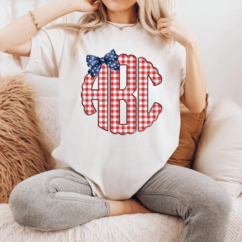 Op de afbeelding: Wit T-shirt met een rood-wit geruit monogram met de letters "ABC". Een blauwe strik met witte sterren siert de bovenkant van het monogram. Het T-shirt is een casual stijl.