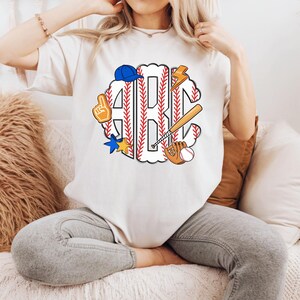 Puede incluir: Camiseta color crema con un dise&ntilde;o tem&aacute;tico de b&eacute;isbol. El dise&ntilde;o incluye las letras "ABC" en un patr&oacute;n de costura de b&eacute;isbol, junto con gr&aacute;ficos relacionados con el b&eacute;isbol, como un bate, un guante, una pelota y una gorra de b&eacute;isbol azul.