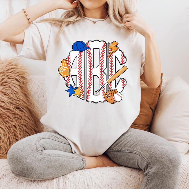 Puede incluir: Camiseta color crema con un dise&ntilde;o tem&aacute;tico de b&eacute;isbol. El dise&ntilde;o incluye las letras "ABC" en un patr&oacute;n de costura de b&eacute;isbol, junto con gr&aacute;ficos relacionados con el b&eacute;isbol, como un bate, un guante, una pelota y una gorra de b&eacute;isbol azul.