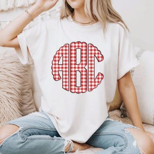 Op de afbeelding: Cr&egrave;mekleurig T-shirt met een rood-wit geruit monogram met de letters "ABC". De letters hebben een afgerond, geschulpt ontwerp. Het shirt is een casual stijl.