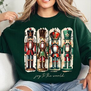 Puede incluir: Sudadera verde con un gráfico de estilo vintage de cuatro soldados cascanueces. El gráfico está en un marco rectangular con fondo blanco y el texto "joy to the world" debajo del marco.