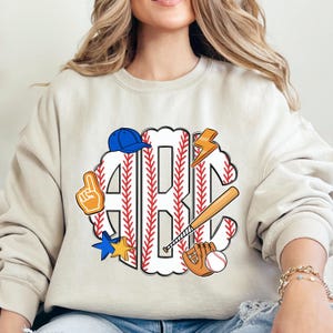 Puede incluir: Sudadera beige con un gr&aacute;fico tem&aacute;tico de b&eacute;isbol. El gr&aacute;fico presenta las letras "ABC" en un dise&ntilde;o de puntada de b&eacute;isbol, rodeadas de iconos relacionados con el b&eacute;isbol como un bate, un guante, una pelota y una gorra azul.