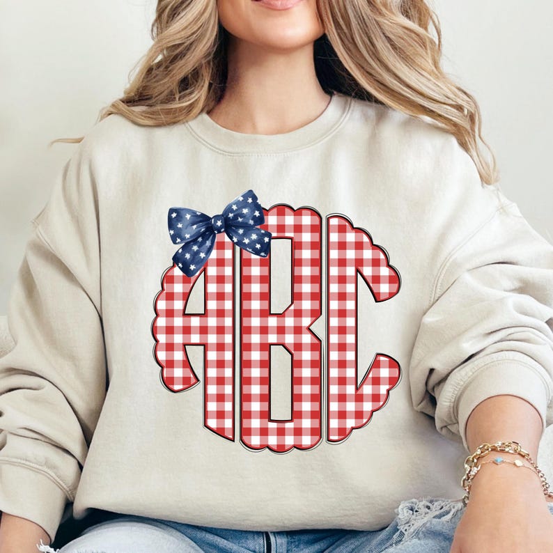 Op de afbeelding: Een beige sweatshirt met een rood-wit geruit monogram met de letters "ABC". Een blauwe strik met witte sterren is boven het monogram geplaatst. De sweatshirt heeft een casual stijl.