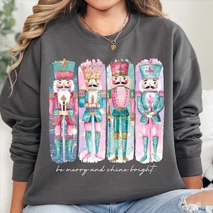 Puede incluir: Una sudadera gris con una ilustración colorida de cuatro soldados cascanueces. El texto "be merry and shine bright." está impreso debajo de la ilustración.
