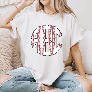 Puede incluir: Camiseta color crema con un dise&ntilde;o tem&aacute;tico de b&eacute;isbol. El dise&ntilde;o incluye las letras "ABC" en un patr&oacute;n de costura de b&eacute;isbol, con costuras rojas y un fondo blanco. La camiseta es de estilo casual.