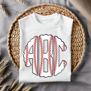 Puede incluir: Camiseta blanca con un dise&ntilde;o de monograma con tem&aacute;tica de b&eacute;isbol. Las letras "ABC" est&aacute;n delineadas en negro con costuras rojas, sobre un fondo blanco en forma de pelota de b&eacute;isbol. La camiseta est&aacute; doblada en una cesta tejida.