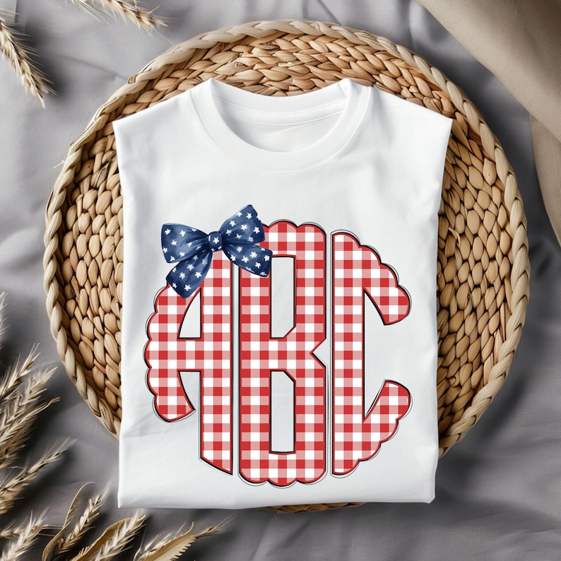 Op de afbeelding: Wit T-shirt met een rood-wit geruit patroon in de vorm van de letters "ABC". Bovenop zit een blauwe strik met witte sterren. Het shirt is opgevouwen in een rieten mand.